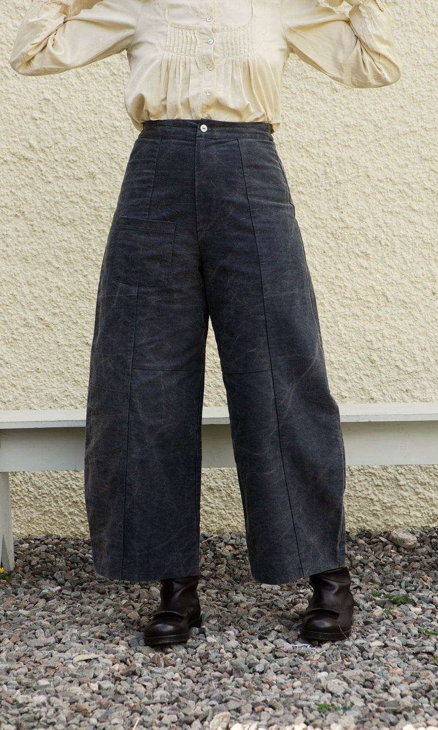 Plümo  Person in Corfe Barrel Pants, cream blouse, and black boots stands by a light wall and white bench.