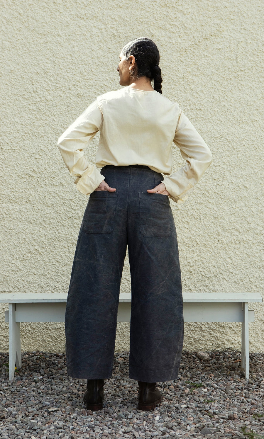 Plümo  Person with braided hair wears Corfe Barrel Pants and cream shirt, facing a pale wall on gravel.