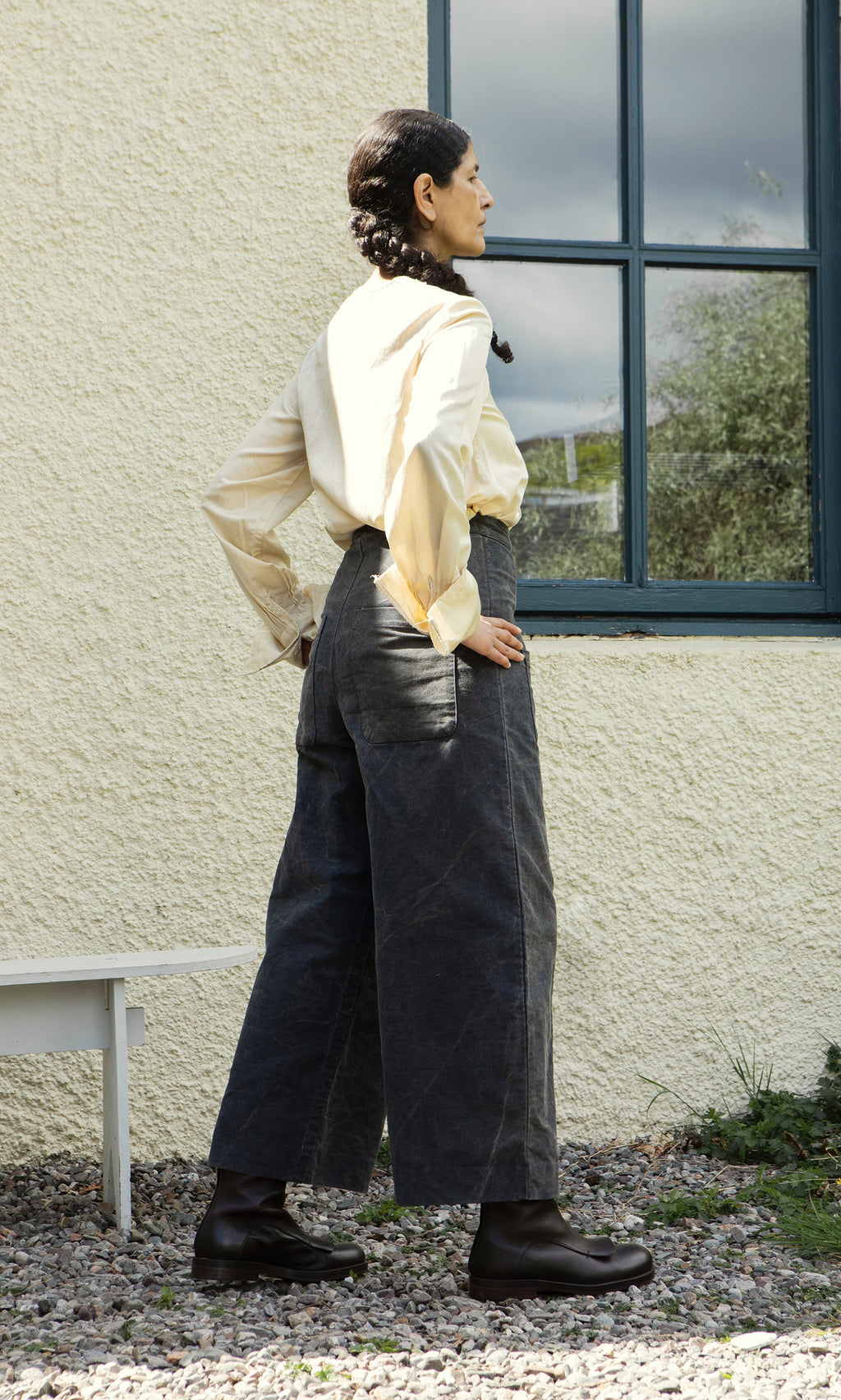 Plümo  A woman in Corfe Barrel Pants and a cream blouse stands outside by a window, hands on hips, facing sideways.