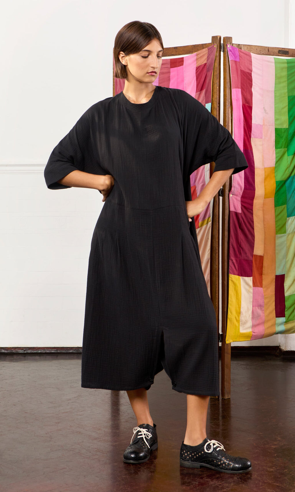 Plümo- Woman in a loose black Culotte Dress and black shoes stands hands on hips before a colorful fabric screen.