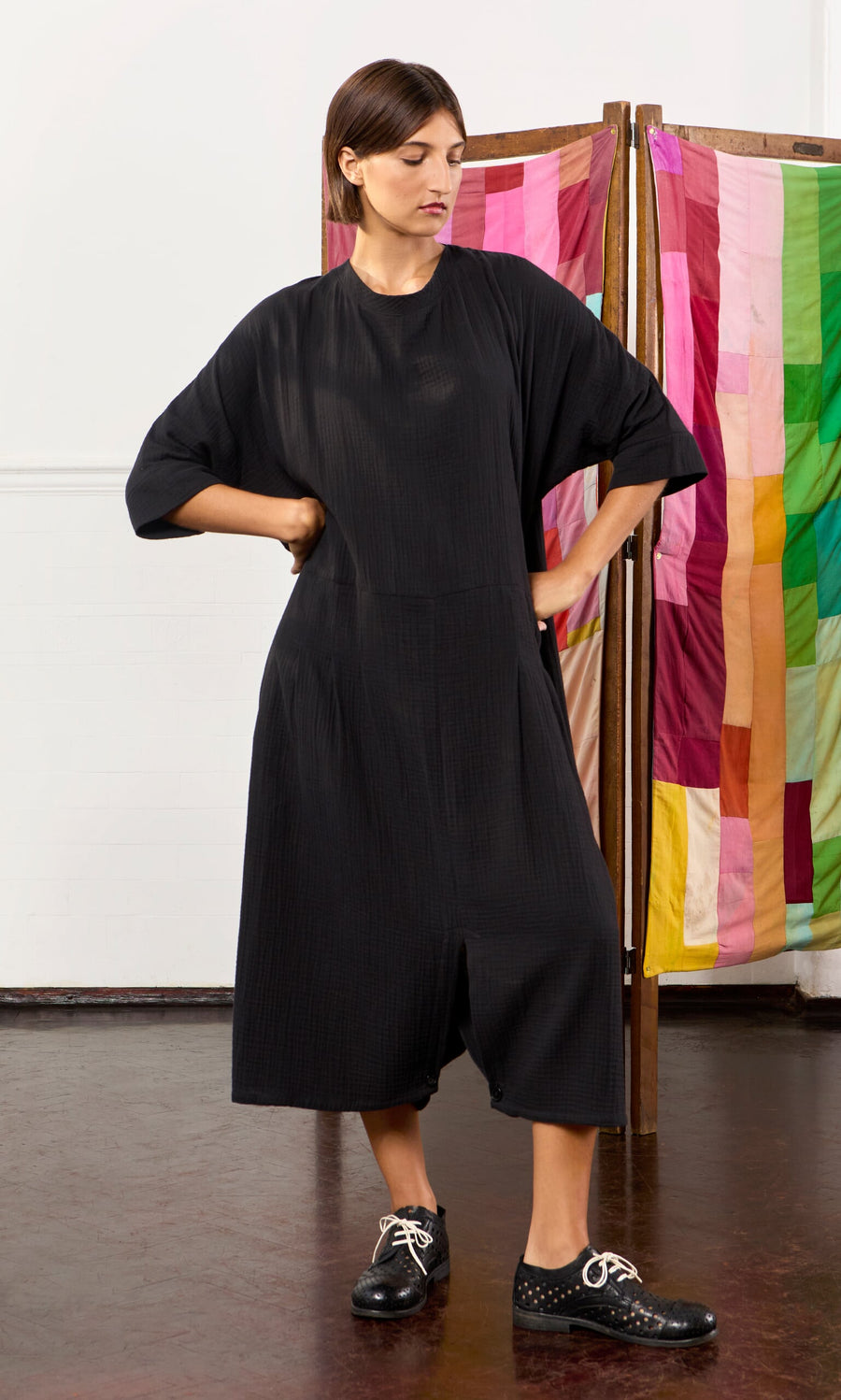Plümo- Woman in a loose black Culotte Dress and black shoes stands hands on hips before a colorful fabric screen.