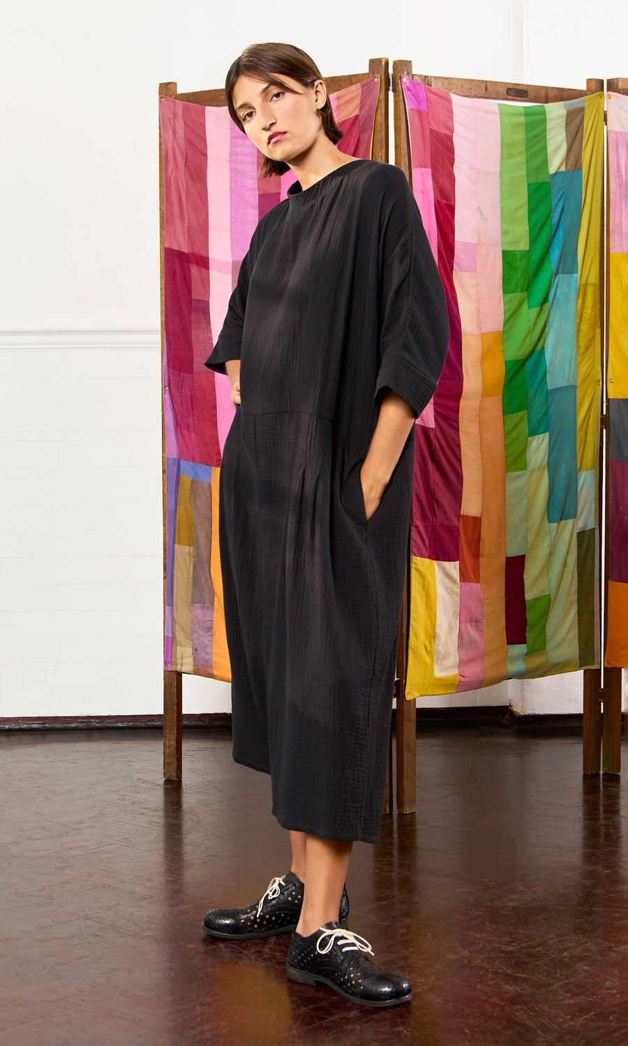 Plümo- Woman in a loose black Culotte Dress and black shoes stands before a colorful patchwork fabric screen.