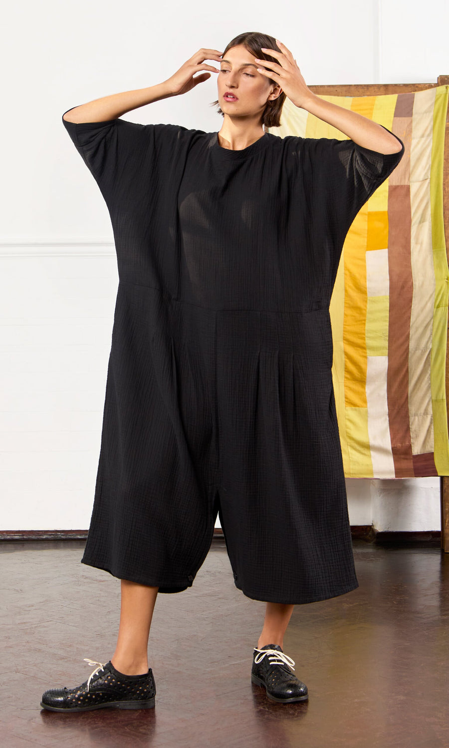 Plümo- Woman in a loose black Culotte Dress stands indoors, adjusting her glasses by a striped fabric backdrop.