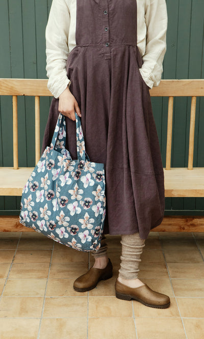 Plümo  Person in a mulberry dungaree dress holds a floral bag, standing on tiled floor by a wooden bench.