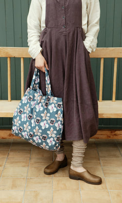 Plümo  Person in a brown dress holds the Dahlia Tote, standing on a tile floor next to a wooden bench.