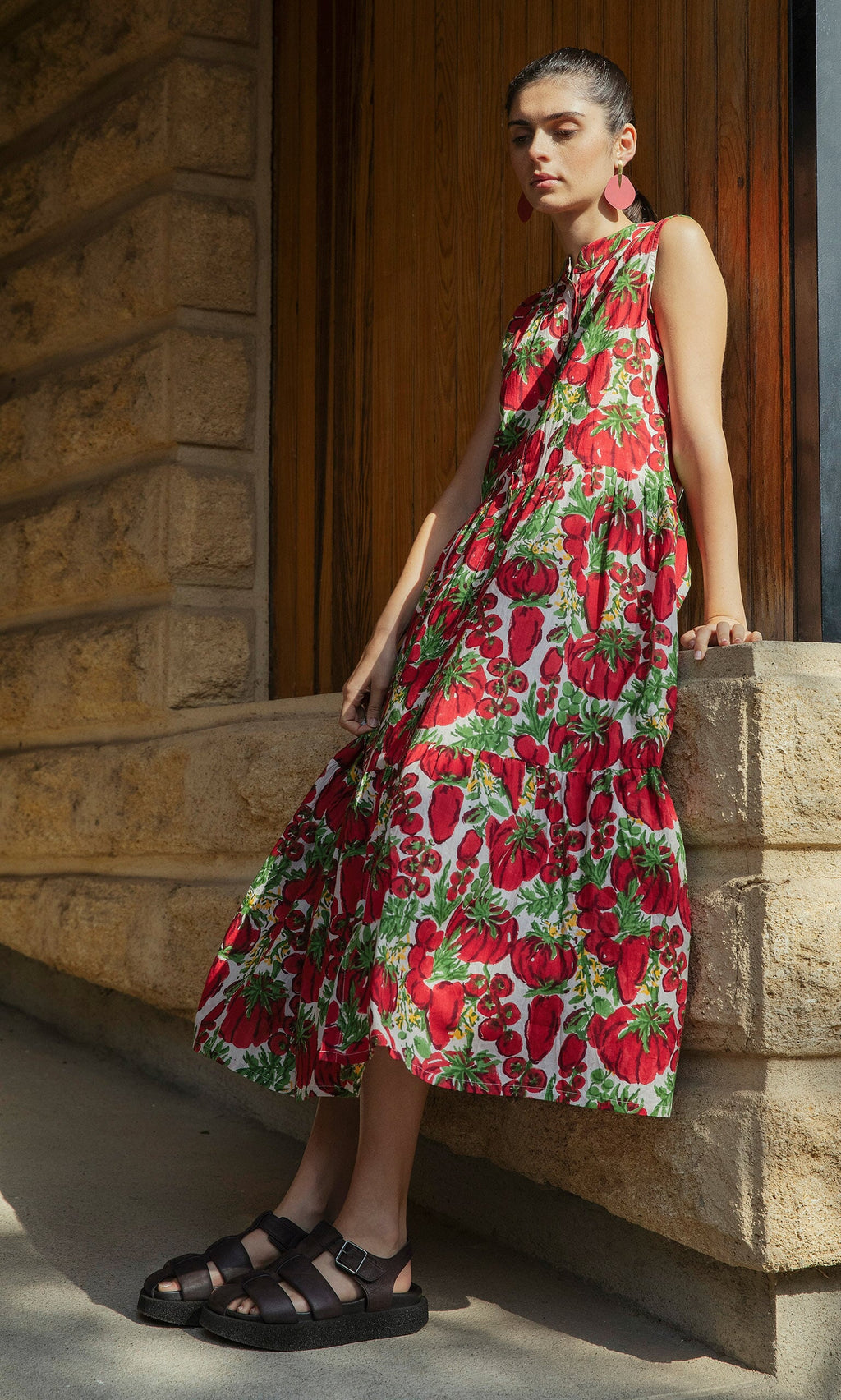 Plümo- Woman in the Datterino Dress—sleeveless cotton-gauze with hand-printed tomatoes—leaning against a stone wall.