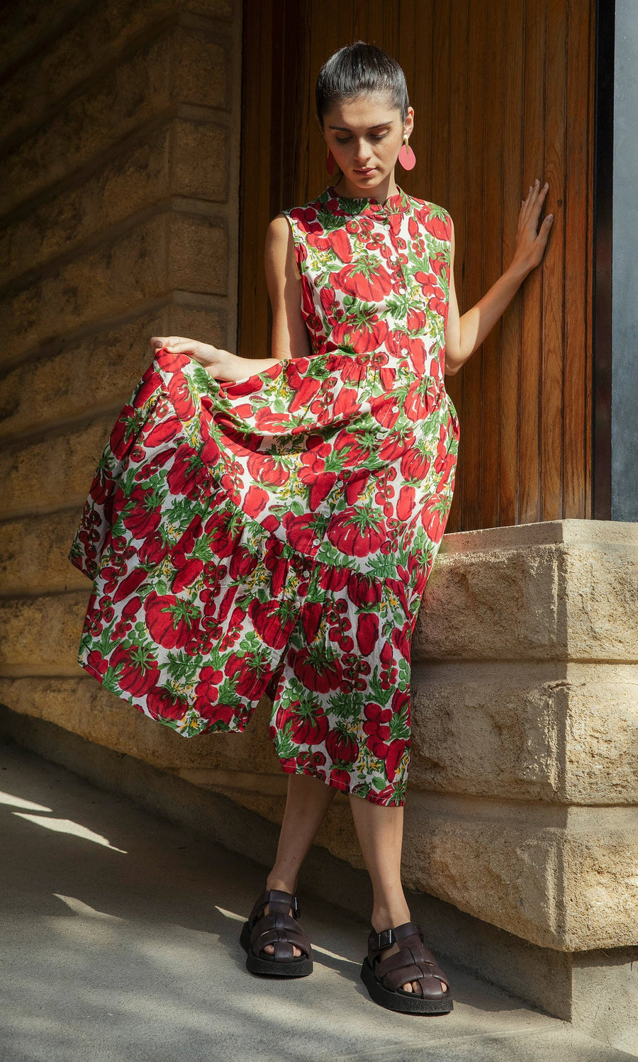 Plümo- Woman in the Datterino Dress with mother of pearl buttons, holding the skirt by a stone wall in sunlight.