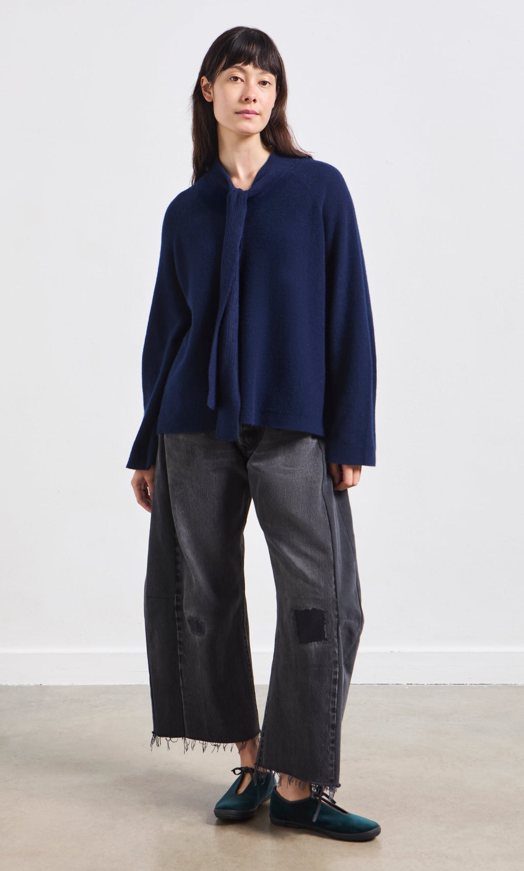 Plümo- Woman stands in a blue Dobbs Jumper with rib knit ties, gray patched jeans, and teal loafers against a plain background.
