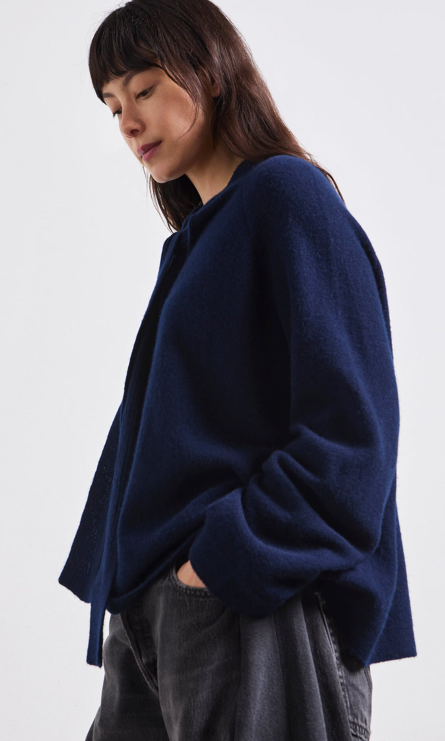 Plümo- Woman with long dark hair in the Dobbs Jumper, a loose navy blue Danish-designed jacket, and black pants, hand in pocket.