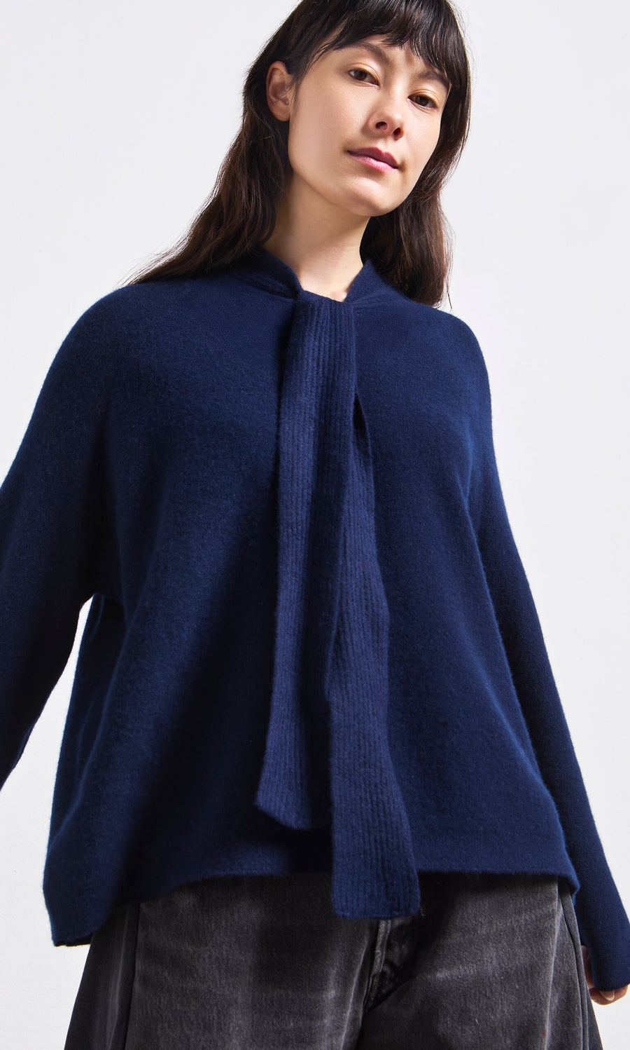 Plümo- Woman wearing the Dobbs Jumper, a Danish design navy sweater with rib knit ties, and dark pants against a light background.