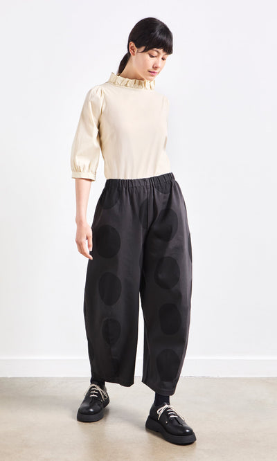 Plümo- Woman in a cream ruffled blouse and Easy Point Pants with large polka dots, standing against a plain white wall.