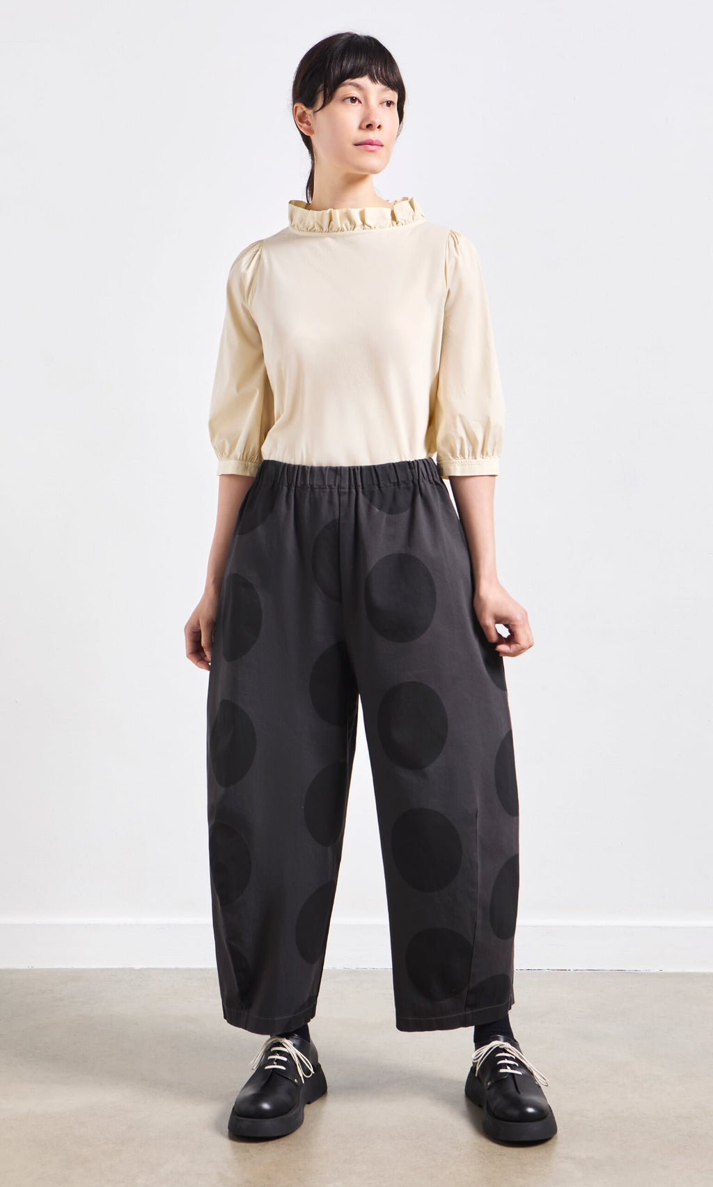 Plümo- Person standing wearing a cream blouse with ruffled collar and Easy Point Pants in large polka-dot cotton-canvas.