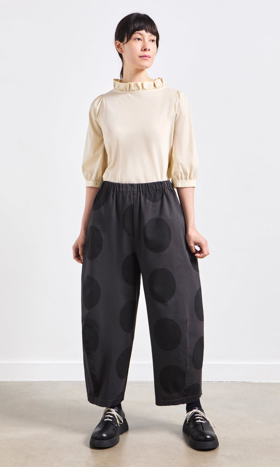 Plümo- Person standing wearing a cream blouse with ruffled collar and Easy Point Pants in large polka-dot cotton-canvas.
