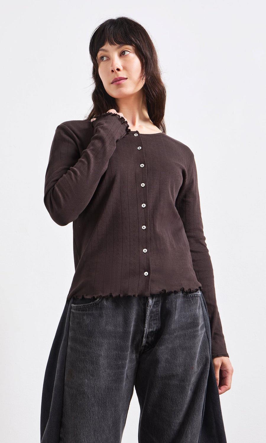 Plümo- Woman wearing the Eddie Cardigan—a dark brown, long-sleeve pointelle knit—paired with black jeans on a white backdrop.