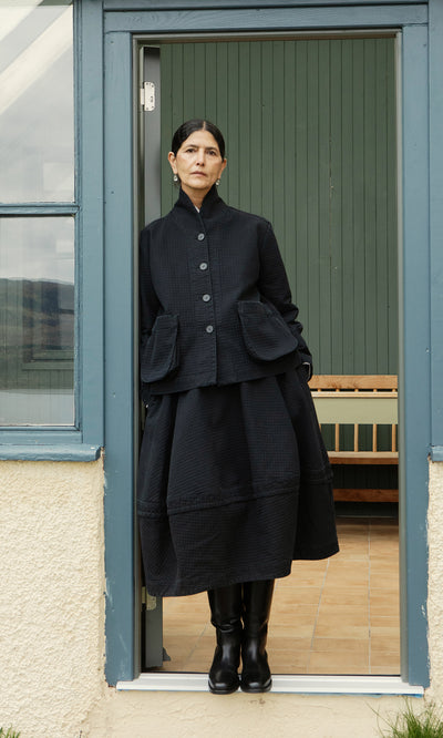 Plümo  Woman wears Rho Skirt - Coal and boots, standing in a doorway with her hands in pockets, green panels behind her.