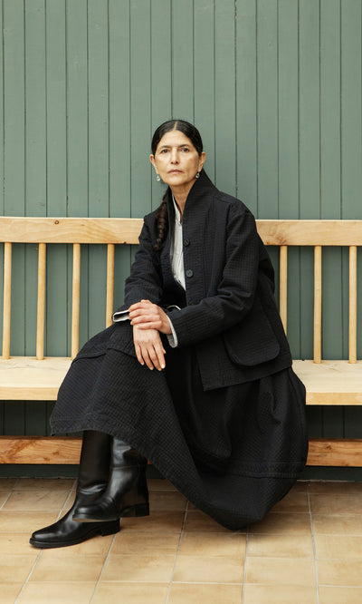 Plümo  Woman in the Envoy Jacket - Coal sits on a bench by a green paneled wall, gazing ahead thoughtfully.