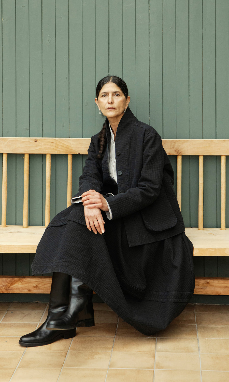 Plümo  Woman in the Rho Skirt - Coal and boots sits on a bench by a green wall, gazing thoughtfully ahead.