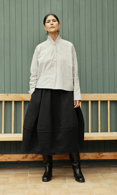 Plümo  A person in a Bennett Blouse - Smoke, black skirt, and boots stands by a green paneled wall and wooden bench.