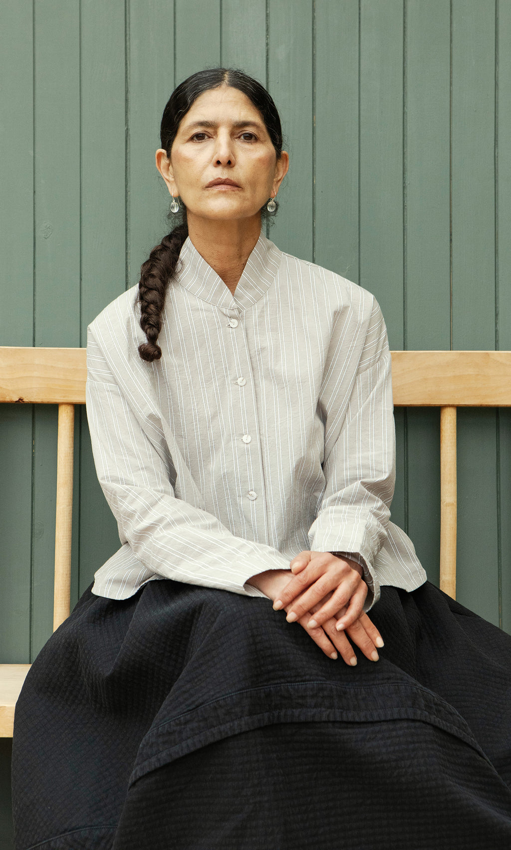 Plümo  A woman with a long braid, striped shirt, and dark skirt sits on a bench wearing Tamsin Earrings.