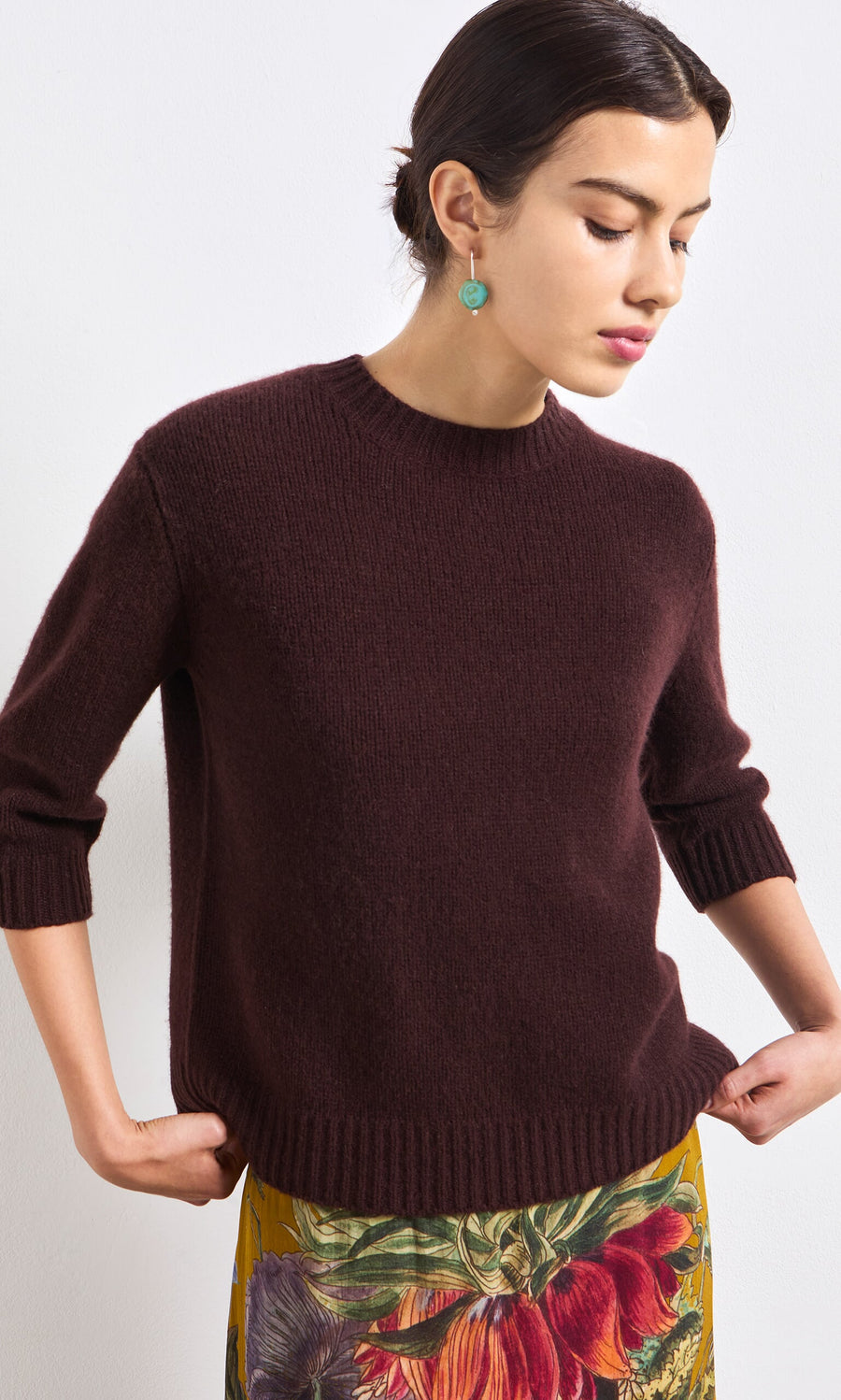 Plümo- Woman in the Bancroft Jumper stands against a white wall, looking down with hands at her sides.