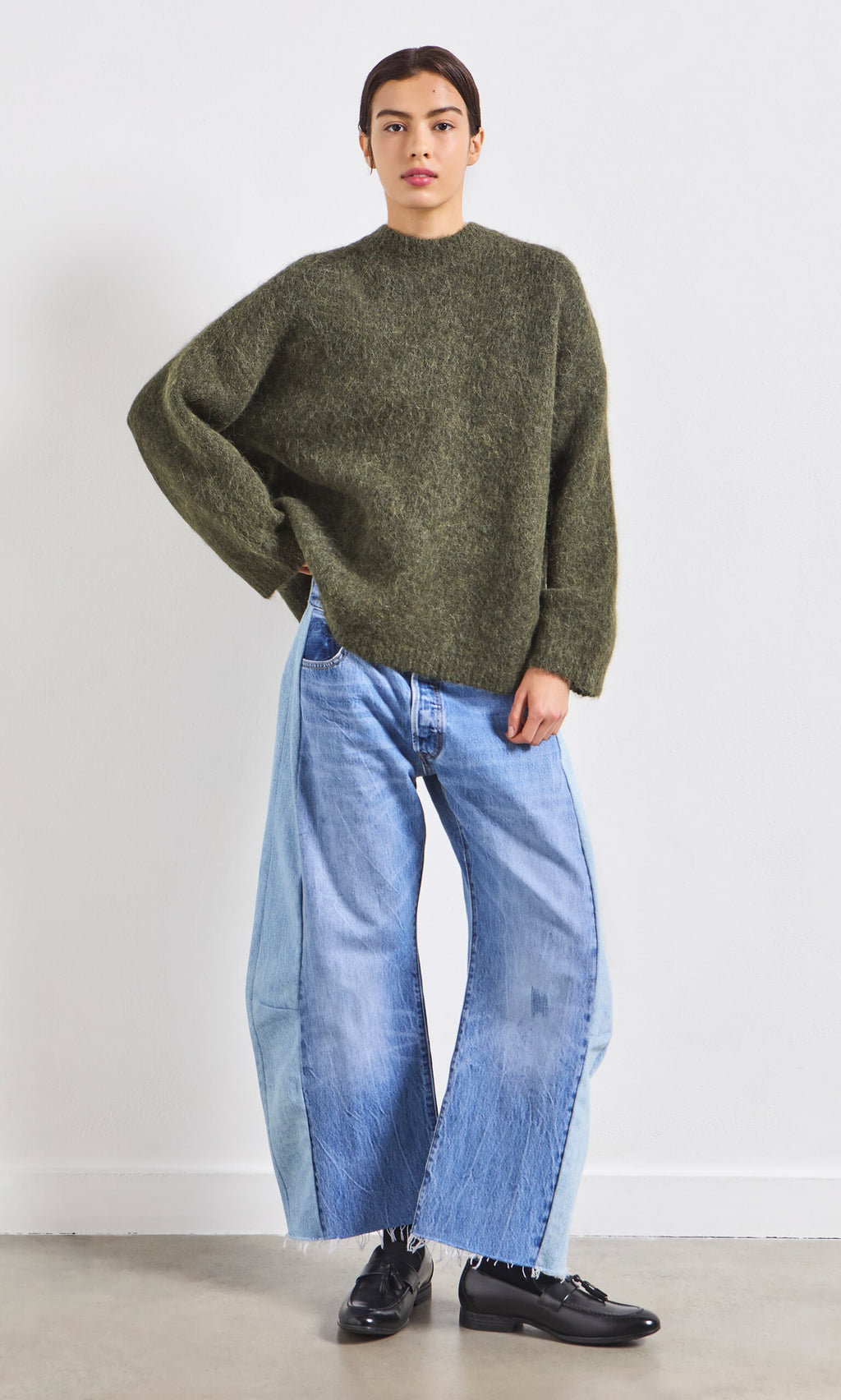 Plümo- Person wears a Fifi Green Jumper and wide-leg light blue jeans, standing against a plain white wall.