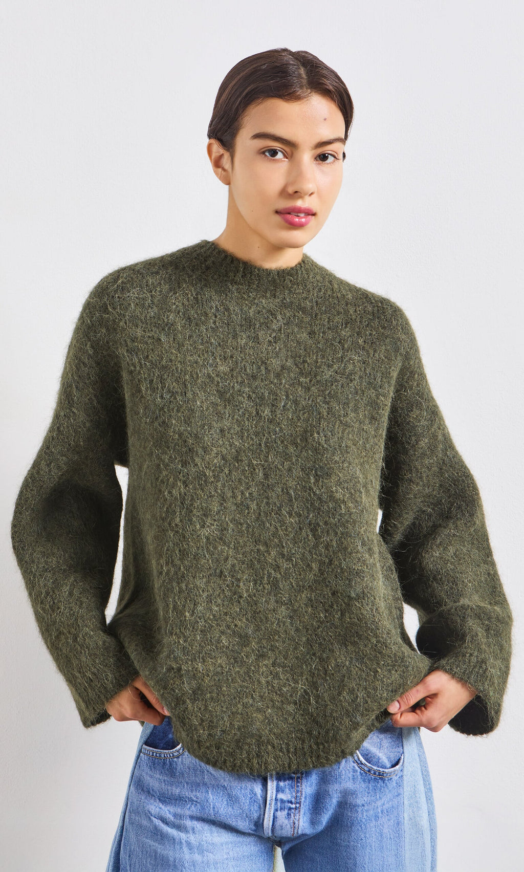 Plümo- Person wears the Fifi Green Jumper and blue jeans, standing with hands in pockets against a white background.