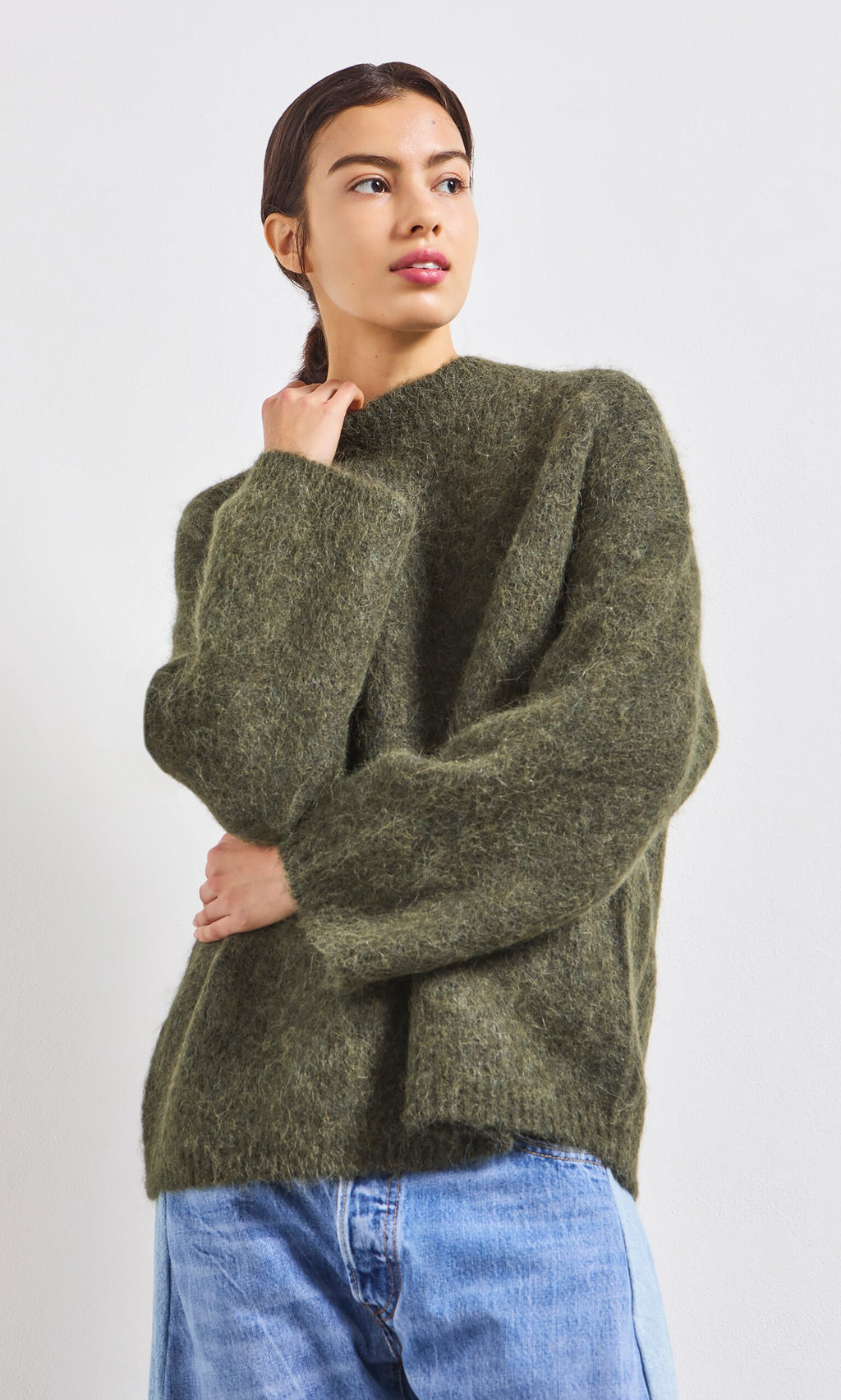 Plümo- Woman wearing the Fifi Green Jumper and blue jeans stands against a white background, looking to the side.