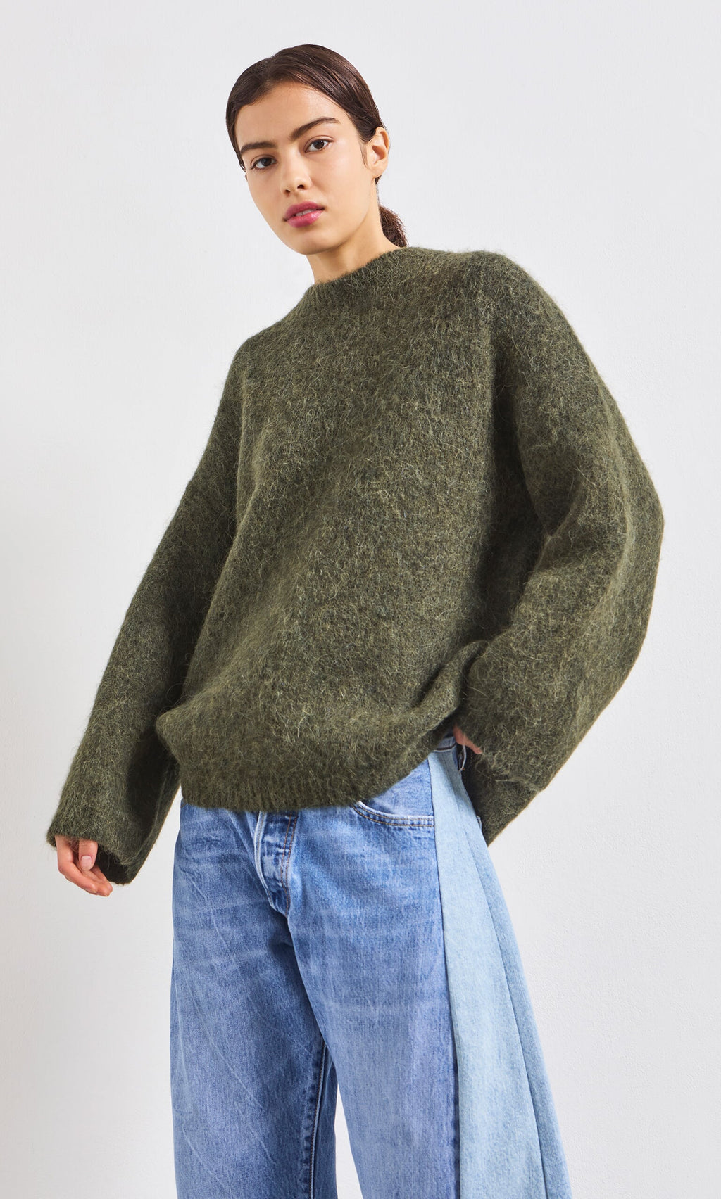 Plümo- Person in a Fifi Green Jumper and two-tone blue jeans stands against a plain white background.