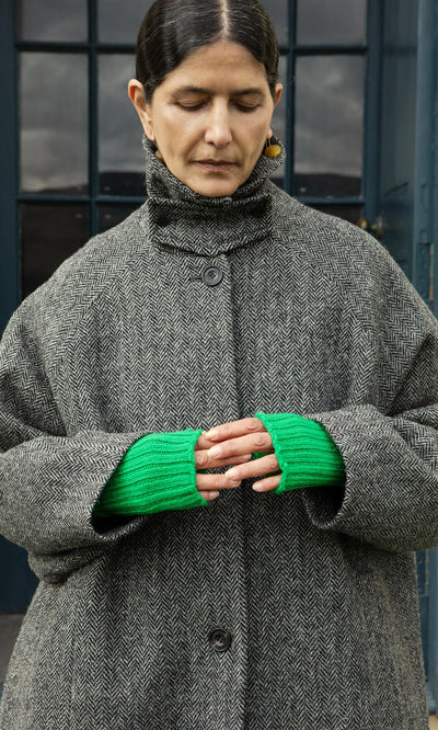 Plümo  A woman in a gray herringbone coat wears Green Wristwarmers, standing outdoors with eyes closed and hands clasped.