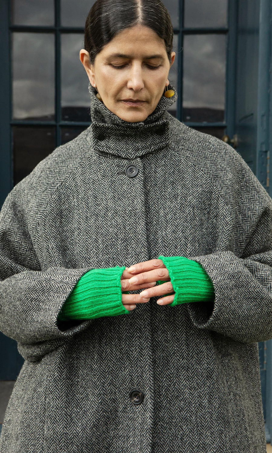 Plümo  A woman in a gray herringbone coat wears Green Wristwarmers, standing outdoors with eyes closed and hands clasped.