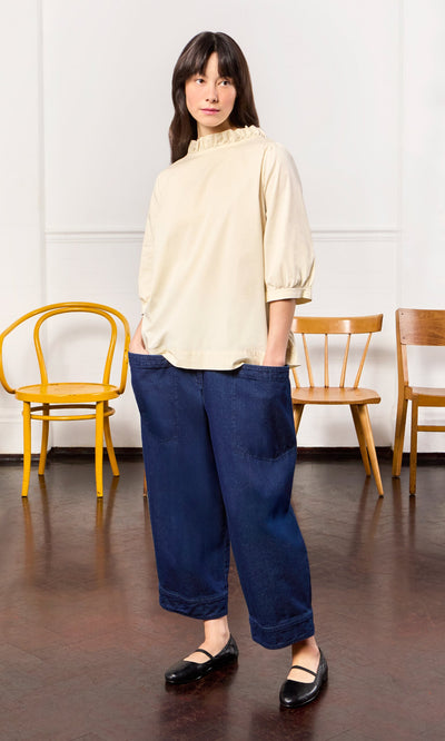 Plümo- Woman wearing the Flint Blouse in ivory cotton with a pie crust frill stands indoors, hands in pockets, chairs behind her.