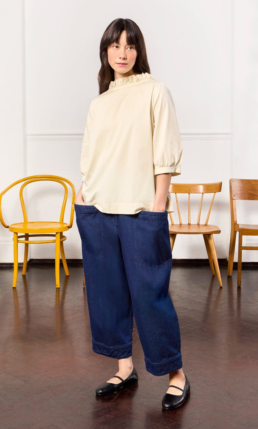 Plümo- Woman wearing the Flint Blouse in ivory cotton with a pie crust frill stands indoors, hands in pockets, chairs behind her.