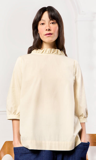 Plümo- Woman wearing an ivory Flint Blouse with pie crust frill and blue pants, standing indoors.