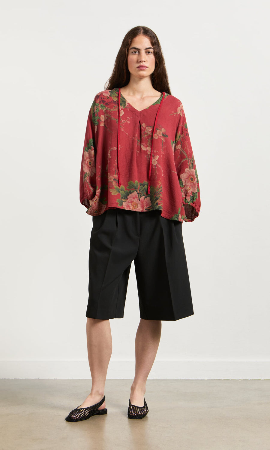 Plümo- A person in a Frida Blouse, black knee-length shorts, and flats stands against a plain background.