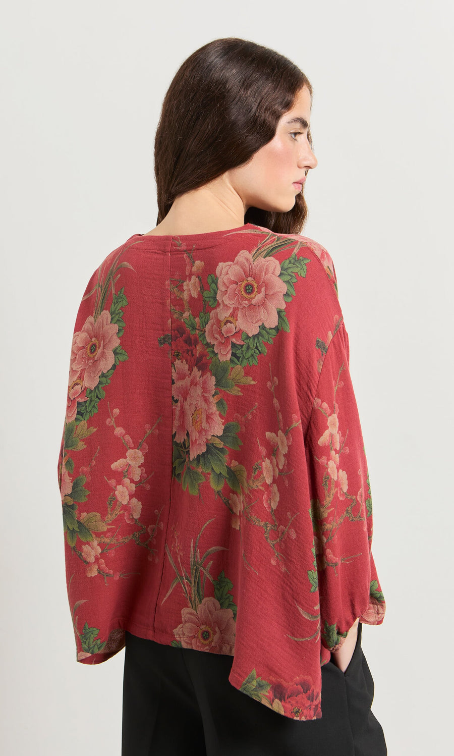 Plümo- Woman with long brown hair wears the Frida Blouse, a floral print linen top, and black pants, facing away.