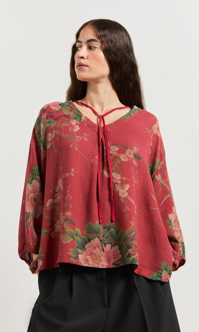 Plümo- Woman wearing the Frida Blouse, a loose red floral linen top with a tie-neck, paired with black pants.