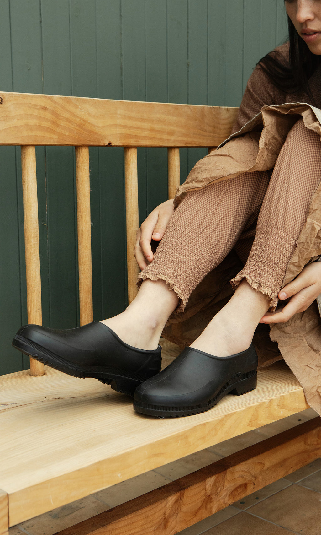 Plümo  Woman in brown on a bench adjusts her pant leg, wearing stylish Gardenia Clogs - Black.