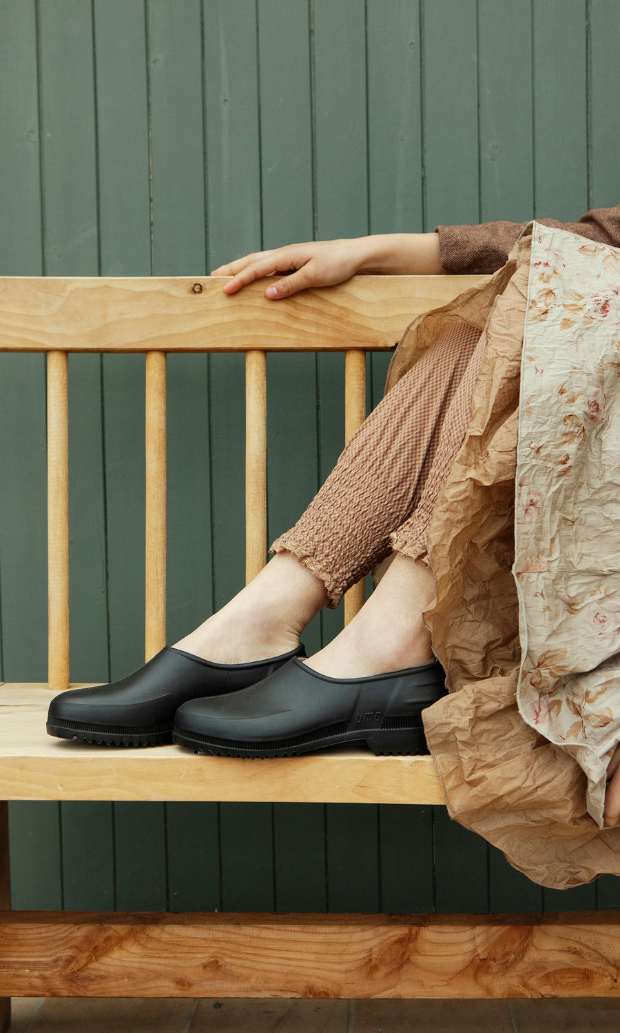 Plümo  Individual on a wooden bench in Gardenia Clogs - Black and textured pants, next to a crumpled floral skirt.
