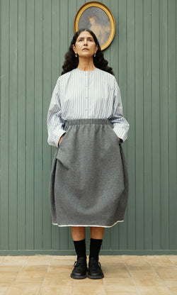 Plümo  Woman in a striped top and Ghent Skirt stands with hands in pockets, gazing up at a portrait above her.