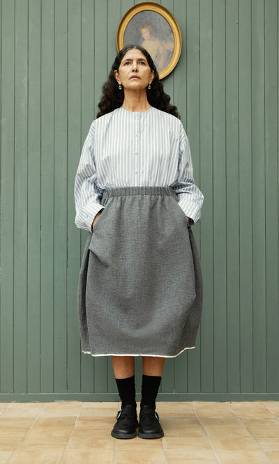 Plümo  Woman in a striped top and Ghent Skirt stands with hands in pockets, gazing up at a portrait above her.
