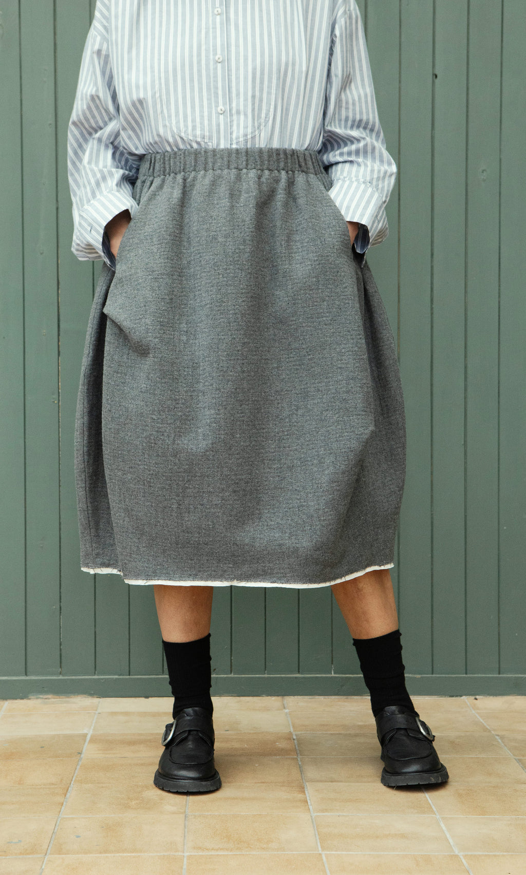 Plümo  Person in a Ghent Skirt, striped shirt, black socks, and sandals stands by a green wooden wall.