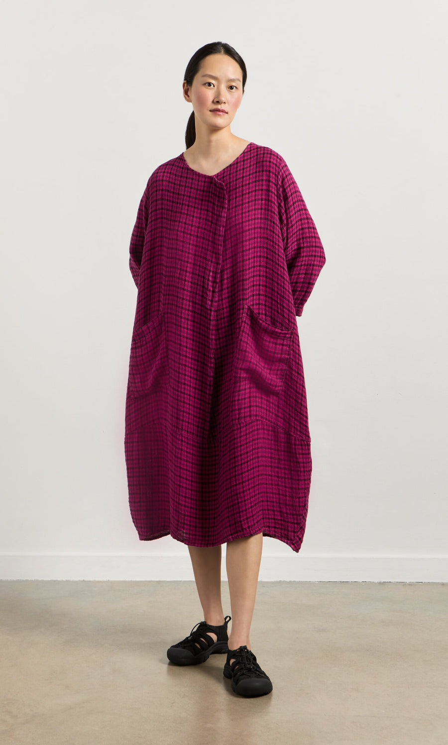 Plümo- Woman in the Gilda Check Dress—an oversized, long-sleeve magenta dress with pockets—stands against a white wall.