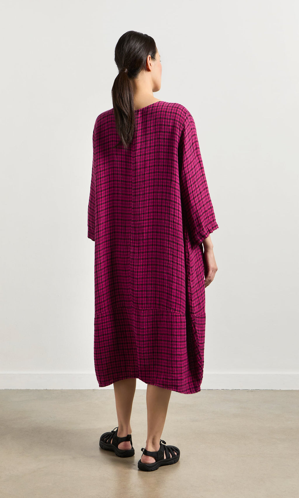 Plümo- Woman faces away in the Gilda Check Dress, an oversized magenta windowpane linen style, paired with black sandals.
