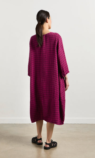 Plümo- Woman faces away in the Gilda Check Dress, an oversized magenta windowpane linen style, paired with black sandals.
