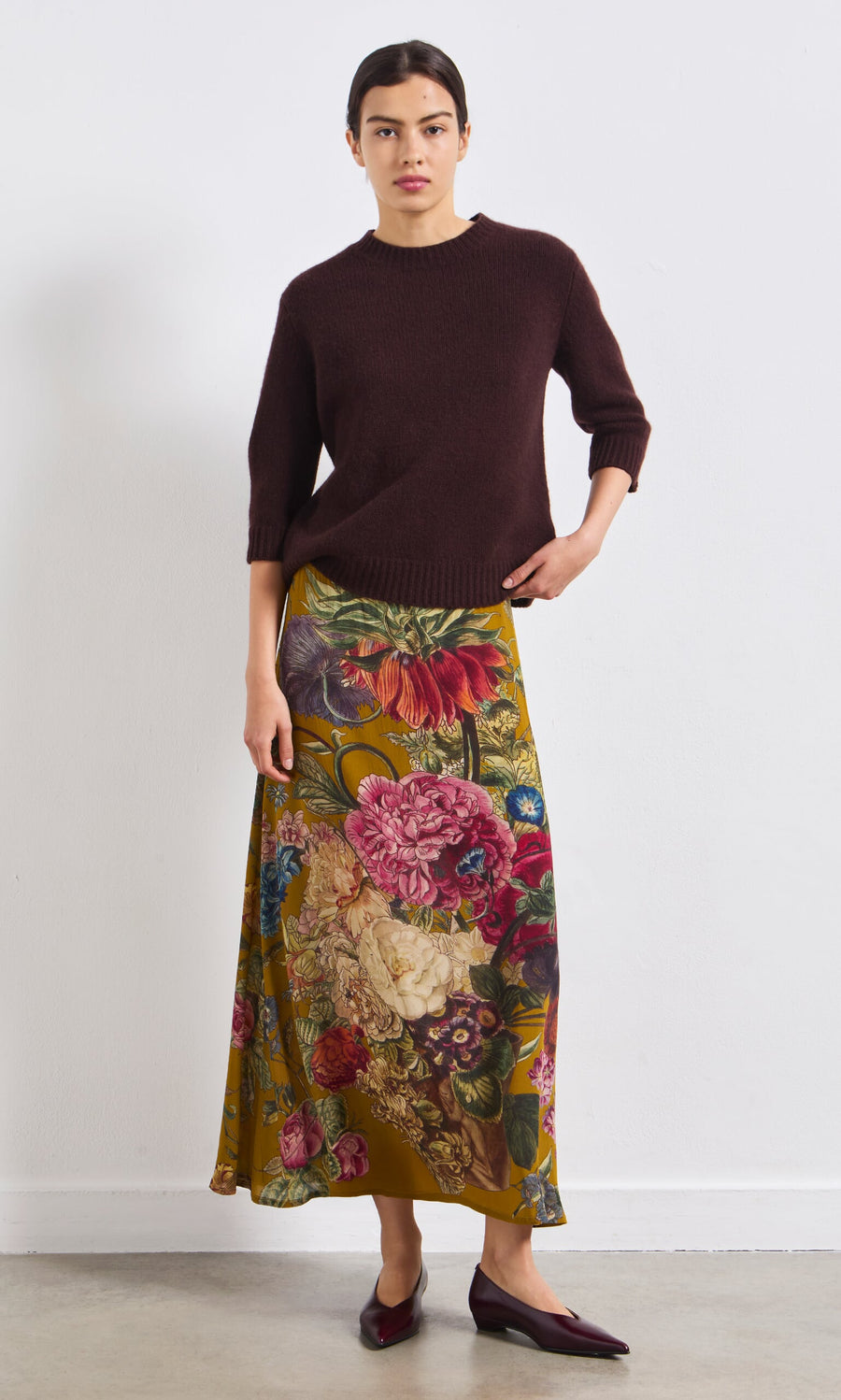 Plümo- A woman in a vintage-inspired dark brown sweater, Golda Skirt, and burgundy flats stands by a white wall.
