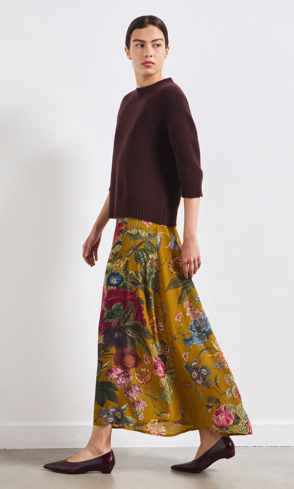 Plümo- Someone wearing a dark sweater and the Golda Skirt walks in burgundy flats against a plain white wall.