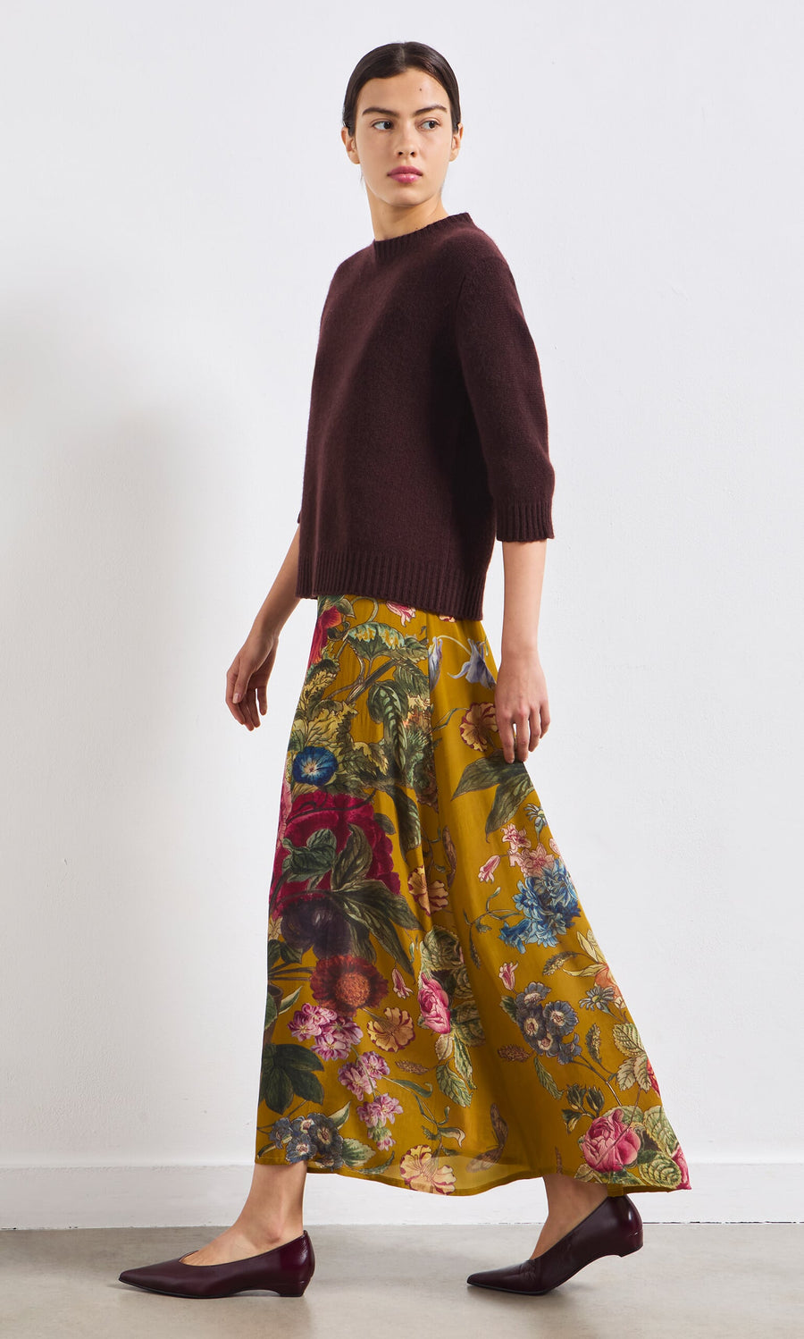 Plümo- Someone wearing a dark sweater and the Golda Skirt walks in burgundy flats against a plain white wall.