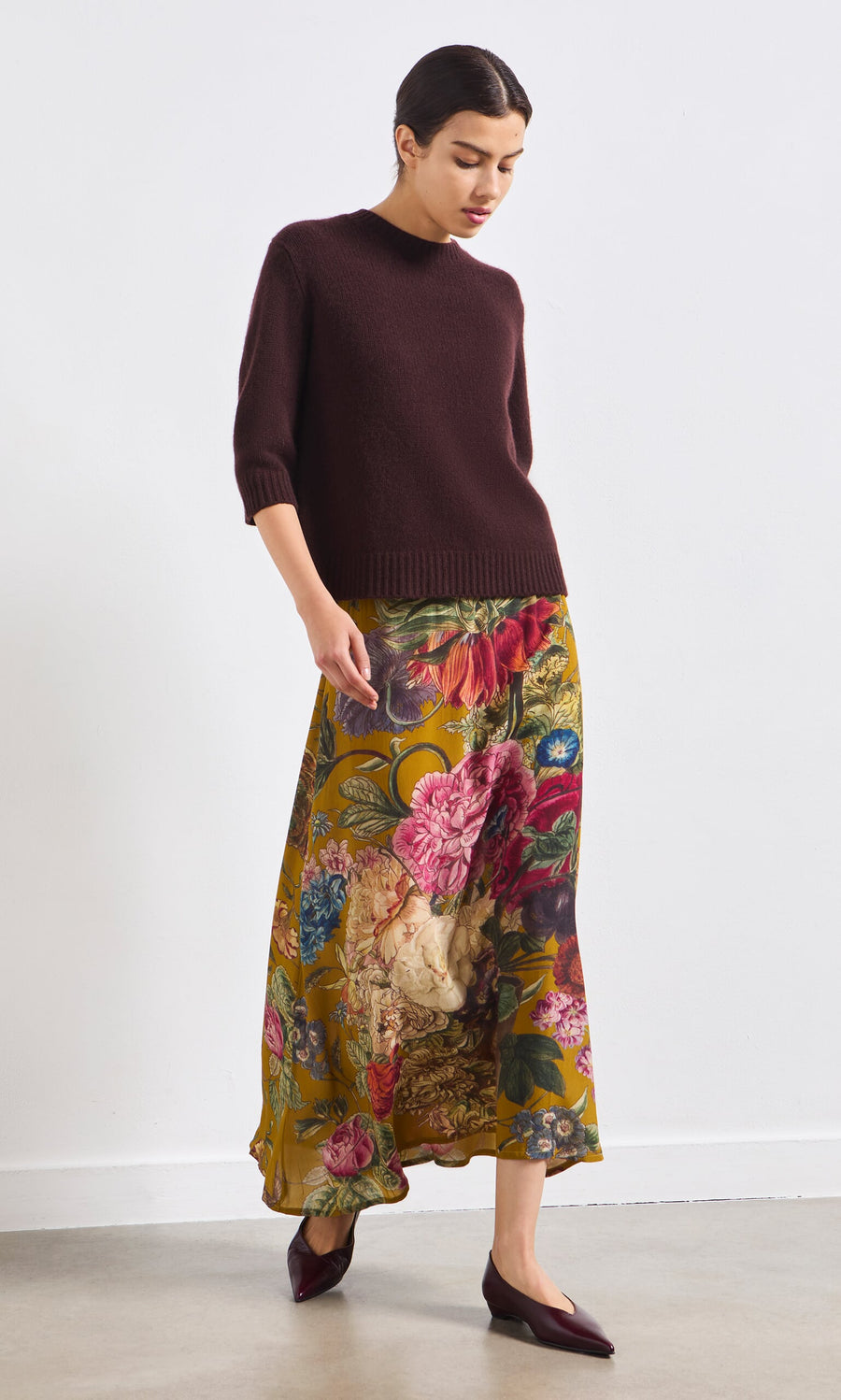 Plümo- Woman wearing a vintage inspired burgundy sweater, Golda Skirt, and burgundy flats stands against a white wall.