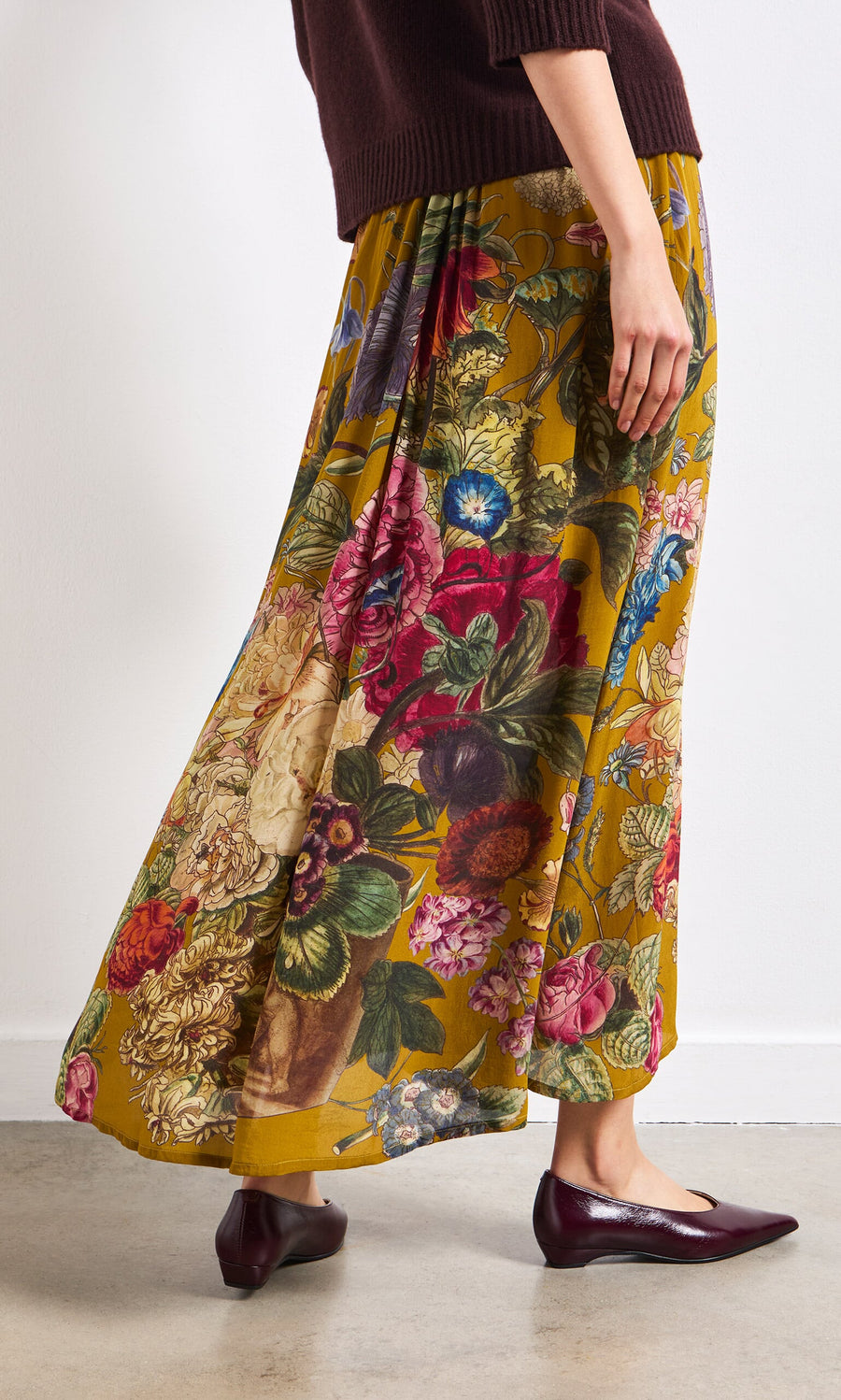 Plümo- Wearing the Golda Skirt, burgundy flats, and a brown sweater, standing indoors.