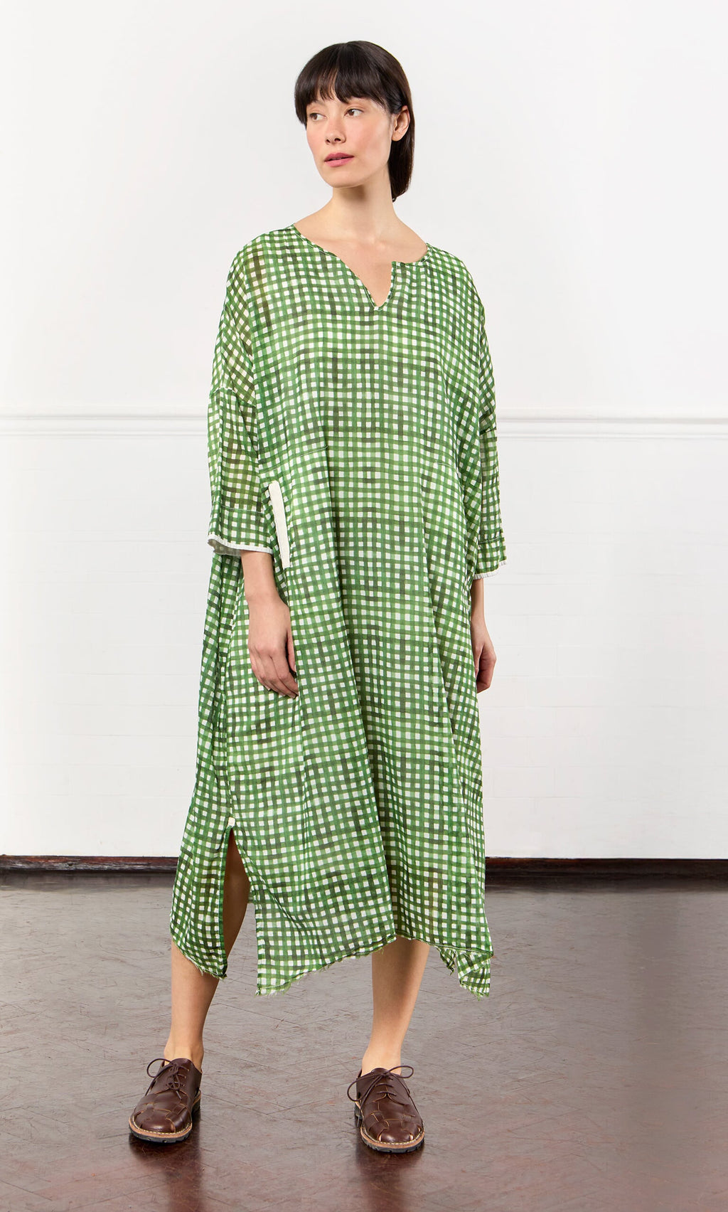 Plümo- Woman in a green gingham dress with brown shoes, standing indoors against a white wall.